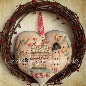 May include: A heart-shaped fabric ornament featuring a painted scene of two houses, a flower pot with red hearts, and the word "LOVE". The ornament is hanging from a wreath of brown twigs with red berries.