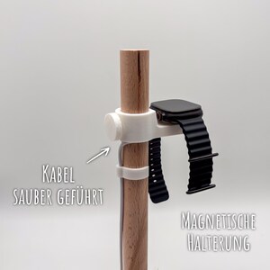 Pode incluir: Um suporte de relógio de madeira com um suporte magnético branco. O suporte apresenta um smartwatch preto com uma pulseira preta. O texto no suporte diz "KABEL SAUBER GEFÜHRT" e "MAGNETISCHE HALTERUNG".
