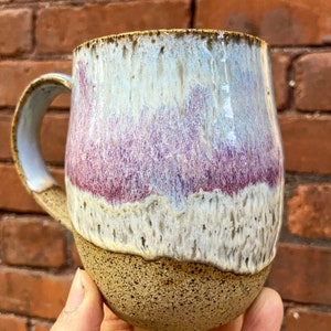 Op de afbeelding: Een handgemaakte keramische mok met een uniek glazuur. De mok heeft een gespikkelde bruine basis, die overgaat in een witte, paarse en blauwe bovenkant. Het handvat is lichtblauw. Perfect voor koffie of thee.