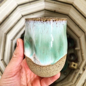 Peut inclure: Une tasse en céramique vert clair avec un bord et une base brun tachetés. La tasse a une forme unique, légèrement irrégulière.