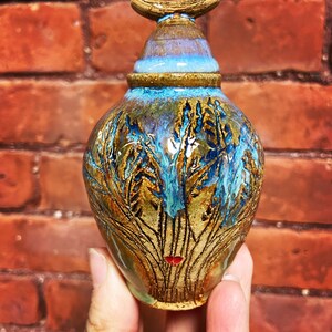 May include: A small, hand-crafted ceramic vase with a blue and brown glaze. The vase has a unique design with a tree-like pattern and a red accent.