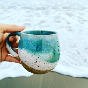 Könnte beinhalten: Eine Keramiktasse mit blauer, weißer und brauner Glasur. Die Tasse hat eine abgerundete Form und eine strukturierte Oberfläche. Sie wird in einer Hand vor einem Hintergrund eines Strandes mit Wellen gehalten.