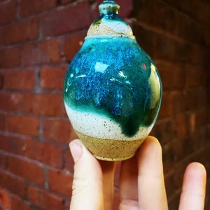 Puede incluir: Un pequeño jarrón de cerámica hecho a mano con un esmalte turquesa, blanco y marrón. El jarrón tiene un cuerpo redondeado y un cuello estrecho.