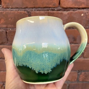 May include: A ceramic mug with a green, blue, and yellow glaze. The mug has a rounded body and a handle.