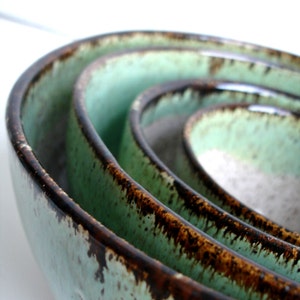 May include: A set of four nesting bowls in varying sizes. The bowls have a light green glaze with a dark brown rim. The bowls are stacked inside each other, showcasing their different sizes and shapes. The bowls are made of ceramic.