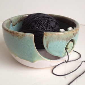 May include: A ceramic yarn bowl with a teal glaze and a white rim. A black ball of yarn is resting in the bowl, with a length of yarn hanging out of the bowl.