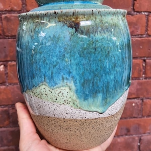 Puede incluir: Un jarrón de cerámica hecho a mano con un esmalte turquesa y azul. El jarrón tiene una base moteada de color canela y una banda blanca y verde. El borde superior es de un turquesa más oscuro. El jarrón se sostiene en una mano.