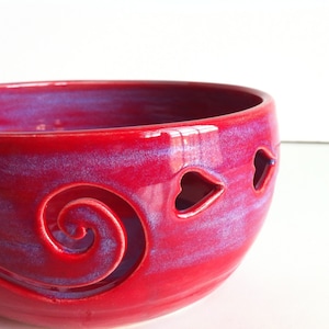 May include: A vibrant red ceramic bowl with a glossy finish. The bowl features a decorative spiral design and heart-shaped cutouts. The interior has a purple hue. This handcrafted bowl is ideal for home decor.