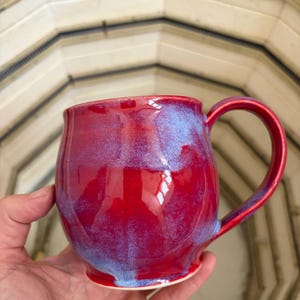 May include: A handmade ceramic mug with a glossy red glaze and blue accents. The mug has a rounded body, a sturdy handle, and a white base. The mug is held in a hand, showcasing its size and shape. The mug is a piece of functional art.