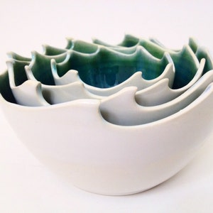 May include: A set of white ceramic bowls with a wave-like design. The interior of the bowls is a gradient of teal and green. The bowls are nested together, showcasing their varying sizes and the depth of the design.