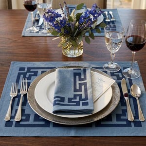 May include: A dining table setting with blue placemats featuring a geometric design. Each place setting includes a white plate, a silver charger, a blue napkin, silverware, and a menu card. Wine glasses and a vase of flowers complete the elegant arrangement.