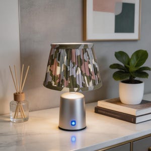 Pode incluir: Candeeiro de mesa prateado com abajur com padrão de camuflagem em tons de verde, rosa e lilás. O candeeiro tem uma luz azul na base. Uma pequena planta em vaso e uma obra de arte abstrata emoldurada estão no fundo. O candeeiro está numa superfície de mármore.