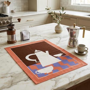 Puede incluir: Escena de cocina con un paño de cocina con un diseño de cafetera y tazas blancas sobre un fondo a cuadros marrón y azul. También se ven una cafetera exprés plateada, una taza y un platillo pequeños y un molinillo de café.