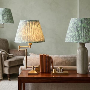 May include: Three lamps with patterned shades in a living room setting. One is a floor lamp, and two are table lamps. The shades have a green and gray design. The table lamps sit on a dark wood table with books and decorative items.