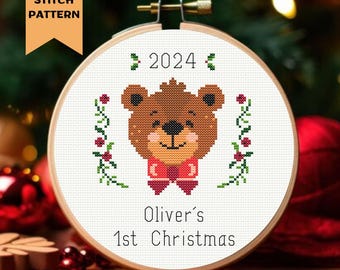 May include: A white cross stitch pattern with a brown bear wearing a red bow tie. The pattern includes the text "2024", "Oliver's 1st Christmas", and a green vine with red berries.