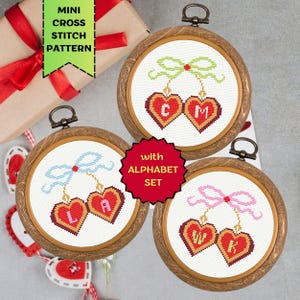 May include: Three round wooden embroidery hoops with cross-stitch heart designs. Each heart has a letter, and is connected to a bow. The text "MINI CROSS STITCH PATTERN" and "with ALPHABET SET" are also visible.