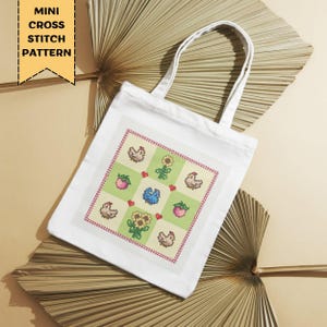 Op de afbeelding: Witte draagtas met een kruissteekpatroon met kippen, zonnebloemen, aardbeien en een blauwe vogel. De tas heeft een rechthoekig ontwerp met een rode rand. De tekst "MINI CROSS STITCH PATTERN" staat in een gele banner.