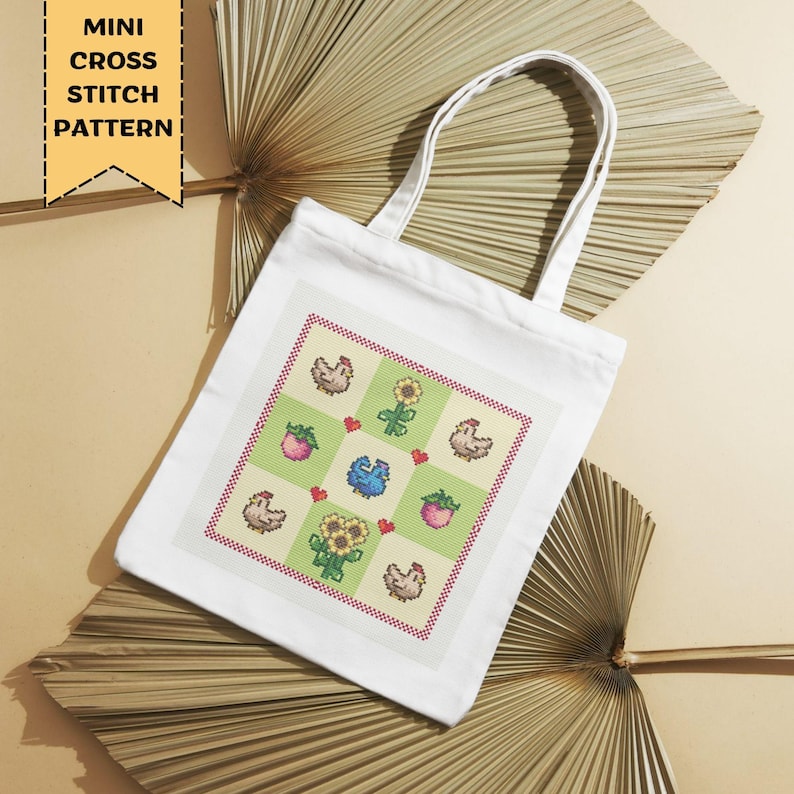 Op de afbeelding: Witte draagtas met een kruissteekpatroon met kippen, zonnebloemen, aardbeien en een blauwe vogel. De tas heeft een rechthoekig ontwerp met een rode rand. De tekst "MINI CROSS STITCH PATTERN" staat in een gele banner.