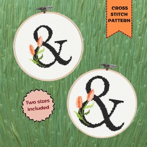 May include: Two circular wooden embroidery hoops containing a cross stitch pattern of a black ampersand symbol decorated with peach-colored tulips and green leaves. An orange banner reads CROSS STITCH PATTERN and a pink badge says Two sizes included.