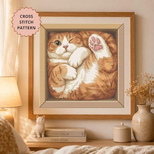 May include: A framed cross stitch pattern featuring an orange and white tabby cat curled inside a square wooden box. The cat has one eye closed in a wink and one paw raised, showing pink paw pads. The text CROSS STITCH PATTERN is in the corner.