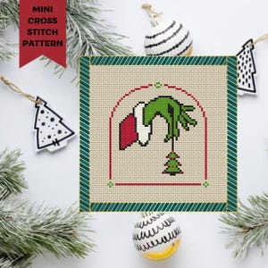 Op de afbeelding: Een kruissteekpatroon met de hand van de Grinch die in een huis reikt en een kerstboom pakt. Het patroon is ingelijst in een groene en gouden rand.