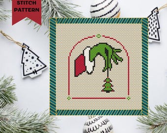 May include: A cross stitch pattern featuring the Grinch's hand reaching into a house and taking a Christmas tree. The pattern is framed in a green and gold border.