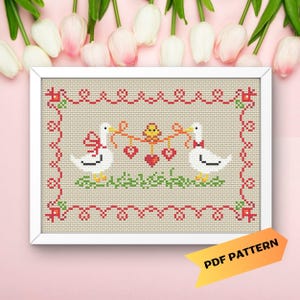 Puede incluir: Patrón de punto de cruz con dos gansos blancos sosteniendo una cuerda de corazones con un pequeño pájaro amarillo en el medio. Los gansos llevan lazos rojos y los corazones son rojos. El patrón está enmarcado en un marco blanco y tiene un borde rojo y blanco.