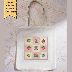 Puede incluir: Un bolso tote blanco con un colorido patr&oacute;n de punto de cruz de varias frutas y flores. El dise&ntilde;o incluye fresas, manzanas y otras im&aacute;genes pixeladas. El texto "MINI CROSS STITCH PATTERN" est&aacute; impreso en una pancarta amarilla.