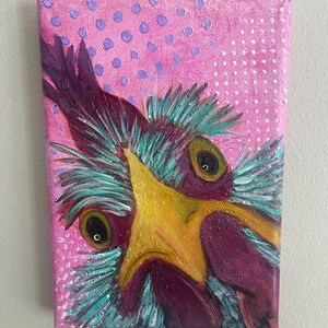 May include: A rectangular canvas painting featuring a close-up of a chicken face with a bright yellow beak, dark purple plumage, and teal-colored feathers. The background is a vibrant pink with purple and white polka dot patterns.