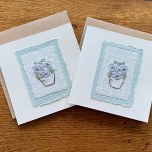May include: Two handmade greeting cards with embroidered floral designs. Each card features a light blue stitched flower pot with purple flowers on a white background, framed by a light blue border. The cards are on a wooden surface.