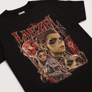 May include: A black short-sleeved t-shirt featuring a graphic print of the character Lae'zel. The design includes the text "LAE'ZEL" in a stylised red font, a red moon with silhouettes of dragons, and detailed illustrations of the character.