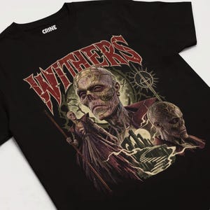 Peut inclure: Un t-shirt noir à manches courtes avec une impression graphique portant le texte "WITHERS" en rouge. Le motif représente trois personnages squelettiques en robes sombres, un château et un symbole solaire sur fond sombre.