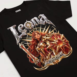 May include: A black t-shirt featuring a graphic print of a female warrior with long red hair, wearing gold armor and holding a sword and a large circular shield. The text "LEONA" appears in large silver letters above, and "SUPPORT" is written below.