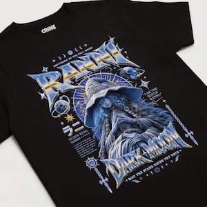 May include: A black t-shirt featuring a graphic print of a blue-toned witch character wearing a wide-brimmed hat. The design includes metallic-style text reading RANNI, DARK MOON, and ELDEN RING, along with celestial symbols, stars, and a moon.