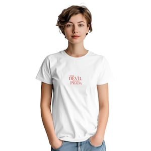 Könnte beinhalten: Weißes T-Shirt mit rotem Text "THE DEVIL WEARS PRADA". Der Text ist mittig auf der Brust platziert. Das Shirt hat kurze Ärmel und einen Rundhalsausschnitt.