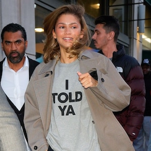 Può includere: Una maglietta grigia con la scritta nera "I TOLD YA". La persona indossa un trench beige. L'immagine è stata scattata all'aperto.