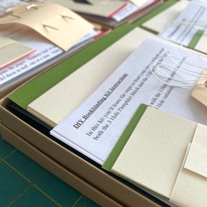 Peut inclure: Gros plan sur des kits de reliure de livres à faire soi-même. Chaque kit contient des instructions, du papier et d'autres matériaux. Les kits sont disposés dans une boîte en carton. Le texte "DIY Bookbinding Kit Instructions" est visible.