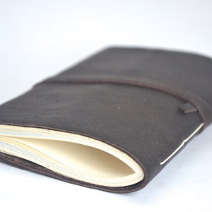 Peut inclure: Un carnet en cuir marron foncé avec une fermeture à lanière enveloppante. Le carnet présente une couverture souple et flexible et contient plusieurs pages de papier couleur crème visibles sur le côté, fixées par une couture blanche.
