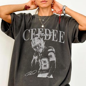 May include: Dark gray t-shirt featuring a grayscale image of a football player in a helmet and jersey. The shirt has the name "CEEDEE Lamb" printed across the top in a distressed font, with the player's signature below.
