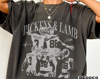 May include: Pepper-colored t-shirt featuring a graphic design with the text "PICKENS & LAMB" and football players. The design includes the numbers 3 and 88, along with signatures. The shirt is a vintage style.