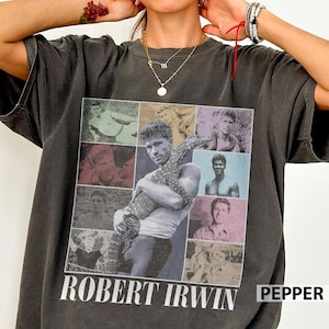 May include: Dark gray t-shirt featuring a collage of images of Robert Irwin, including one with a lizard. The shirt has the name "ROBERT IRWIN" printed in white at the bottom. The word "PEPPER" is printed in white on the right side.