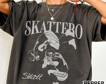 May include: Dark gray t-shirt with the word "SKATTERO" in large, distressed white letters. The shirt features a graphic of football players in action. The word "Slatt" is printed below the graphic.
