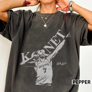May include: Dark gray t-shirt featuring a basketball player graphic with the name "KORNET" and the number 7. The player is wearing a Spurs jersey and has a signature. The word "PEPPER" is at the bottom right.