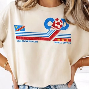 May include: Cream-colored t-shirt featuring a graphic design for Congo DR Soccer. The design includes the flag of the Democratic Republic of the Congo, the text "CONGO DR SOCCER" and "WORLD CUP '26", and a soccer ball.