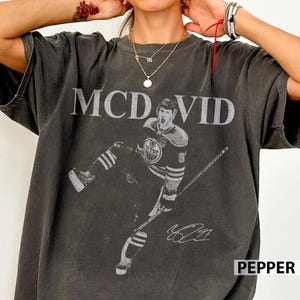 May include: Dark gray t-shirt featuring a graphic of a hockey player with the name "MCDAVID" above the image. The player is wearing a hockey jersey and holding a hockey stick. The shirt also has the word "PEPPER" printed on the bottom right.