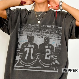 May include: Dark gray t-shirt featuring a hockey-themed graphic. The graphic depicts two hockey players, with the names "CELEBRINI" and "SMITH" and the numbers "71" and "2" on their jerseys. The word "PEPPER" is printed on the bottom right.