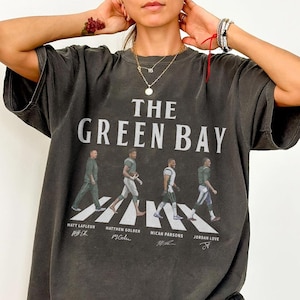 May include: Dark gray t-shirt featuring the text "THE GREEN BAY" above an image of four people walking across a crosswalk. The names Matt LaFleur, Matthew Golden, Micah Parsons, and Jordan Love are printed below the image.