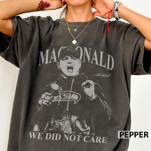 May include: Dark gray t-shirt featuring a graphic of a man with the text "MAC DONALD" above his image and "WE DID NOT CARE" below. The shirt also has the text "PEPPER" in the bottom right corner.