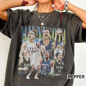 May include: A charcoal gray short-sleeve t-shirt featuring a vintage-style graphic print of Minnesota Lynx basketball players in action. The design includes the text MINNESOTA in large blue letters and various player images with basketballs.