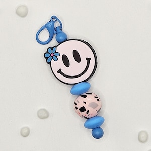 May include: A pink smiley face keychain with a blue flower and black outline. The keychain has a blue clasp and is adorned with blue and pink beads with black speckles. A fun and colorful accessory.